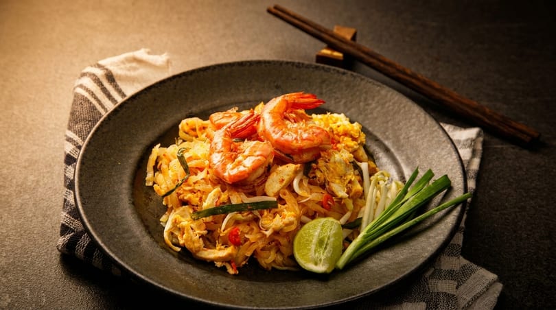 Pad Thai with shrimp, lime, and green onions on a dark plate with chopsticks.