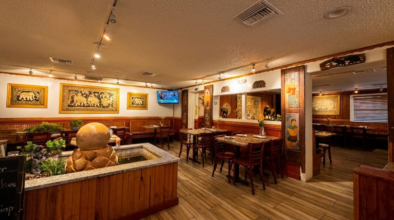 Cozy Thai restaurant interior with wooden tables, chairs, elephant tapestries, and a decorative fountain.
