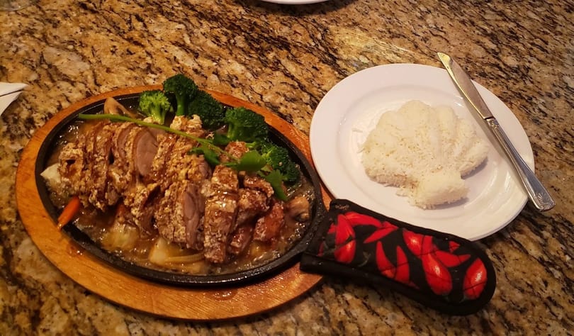 Sizzling platter of sliced duck with broccoli and carrots, served with a side of white rice.