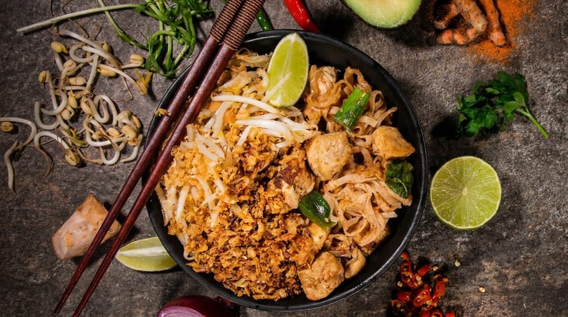Overhead view of chicken Pad Thai with crispy onions, bean sprouts, and lime, surrounded by spices.