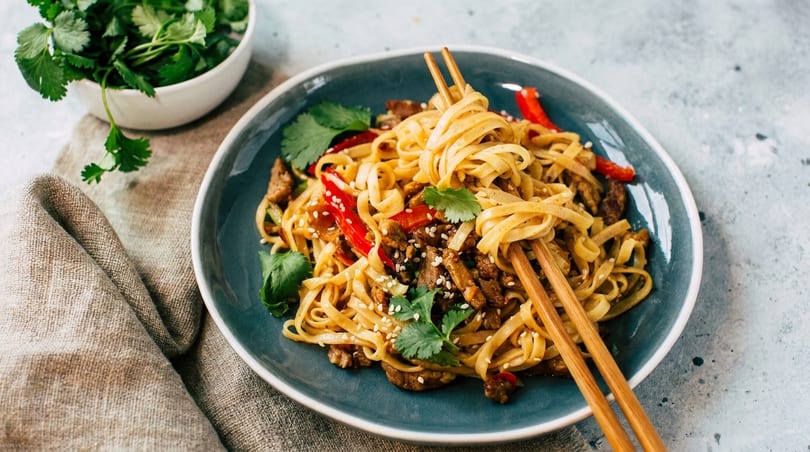 Noodles with beef, red peppers, sesame seeds, and cilantro in a blue bowl with chopsticks.