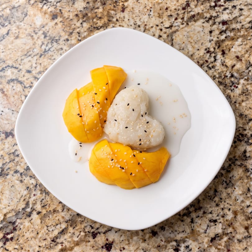 Mango sticky rice with heart-shaped rice, coconut milk, and sesame seeds on a white plate.