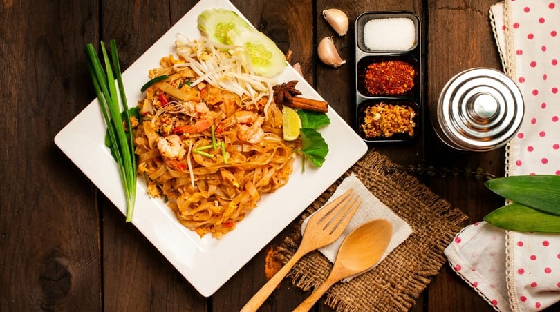 Pad Thai with shrimp, bean sprouts, and lime, served with spices on a wooden table.